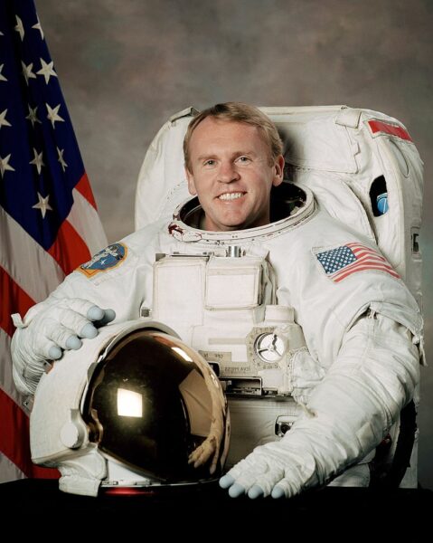a man smiles. He is wearing an astronaut suit in white. He holds a helmet. an american flag is in the background