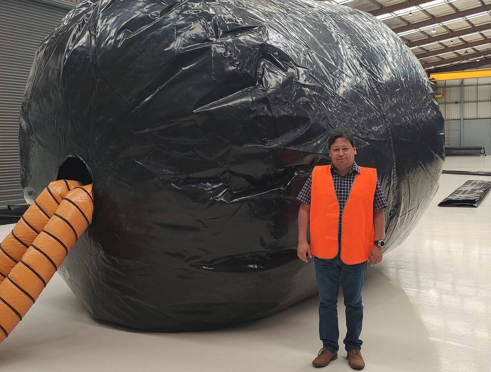 A man stands in from of a giant black inflated ball. The ball has two long orange pipes coming out of it