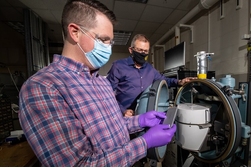 two men in masks in a lab loading a hand-sized battery into a spherical chamber
