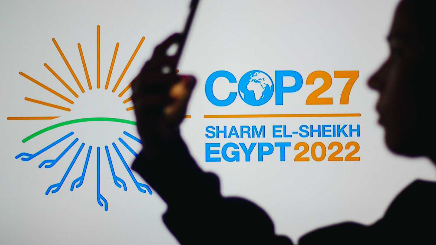 person using a phone in front of sign saying 'COP27: Sharm-el-Sheikh, Egypt'