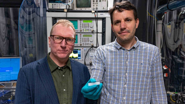 two-scientists-hold-up-photonic-chip-with-computer-stacks-behind