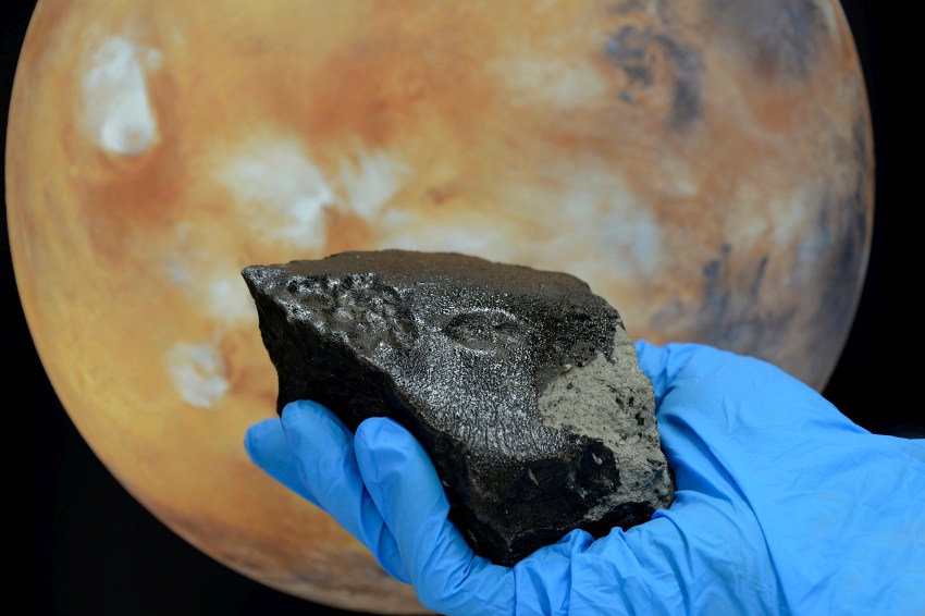 A gloved hand holds a meteorite in front of an image of mars