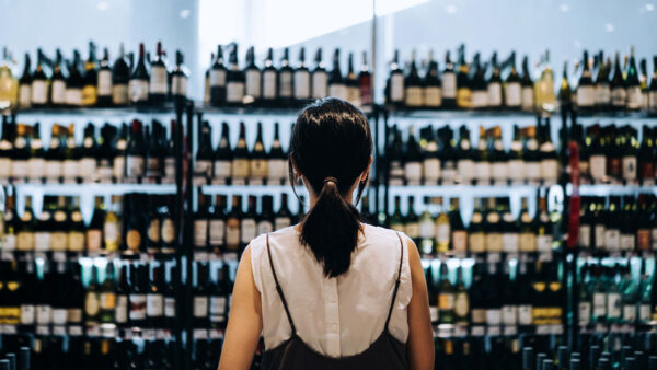 Rear view of young Asian woman grocery shopping for wines in a supermarket. She is standing in front of the liquor aisle and have no idea which wine to choose from