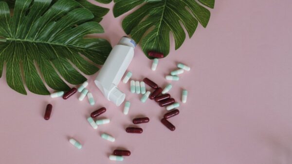 Asthma puffer and vitamin D supplement pills on pink background with leaves